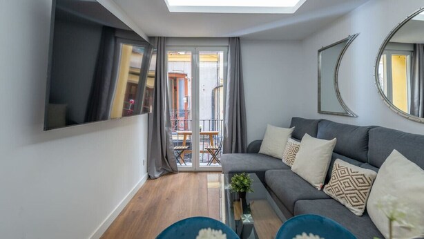 Una sala moderna con un sofá gris, una mesa de centro y un televisor montado en la pared. Se ve un balcón a través de las puertas de vidrio.
