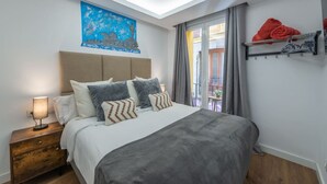 Un dormitorio moderno con tragaluces, una cama con ropa de cama gris, una mesita de noche con lámpara, un cuadro en la pared y un balcón con barandilla.
