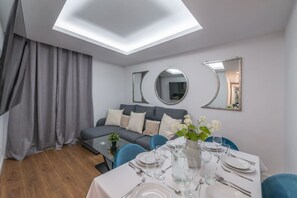 Un salon moderne avec un canapé gris, des fauteuils bleus et une table à manger pour quatre personnes. Deux grands miroirs ornent le mur et une lucarne se trouve au-dessus.