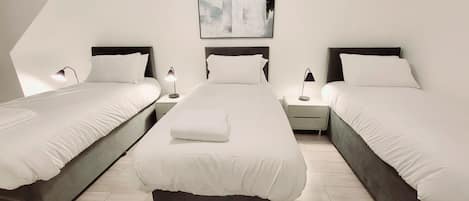 A hotel room with three beds, each with white bedding and dark headboards. There is a painting on the wall above the beds, and two bedside lamps on each nightstand.