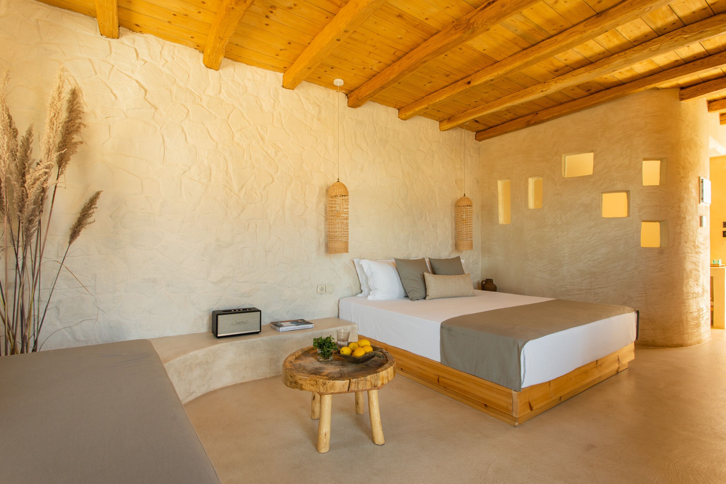 A bedroom with a wooden ceiling, a bed with a grey and white duvet, a bedside table with a radio and a glass of water, and a small round wooden stool with fruits and a plant.