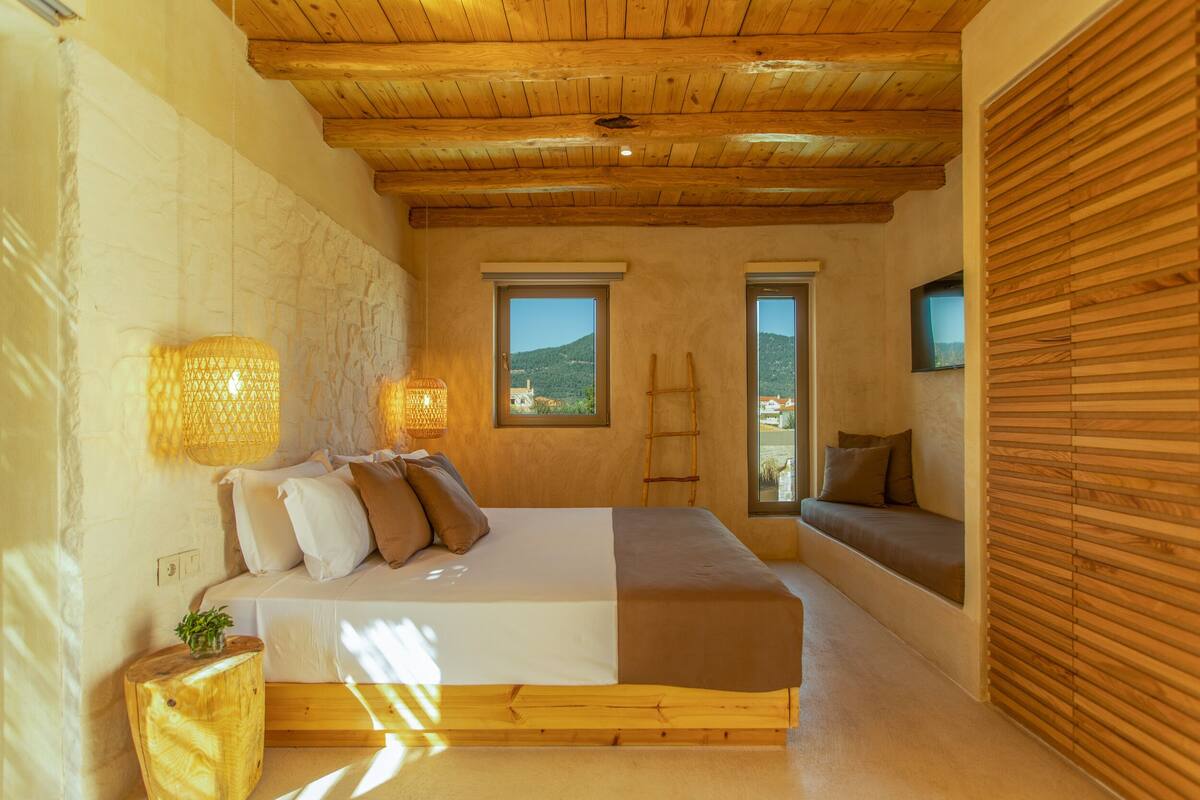 A bedroom with a wooden ceiling, a bed with white and brown bedding, two windows with views of the outdoors, and a wooden bench by the window.