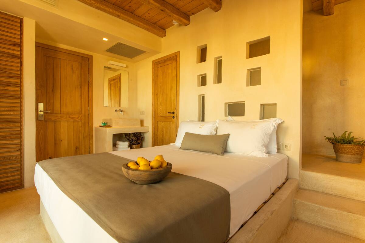 A bedroom with a large bed, a wooden door, a small bedside table, and a potted plant.