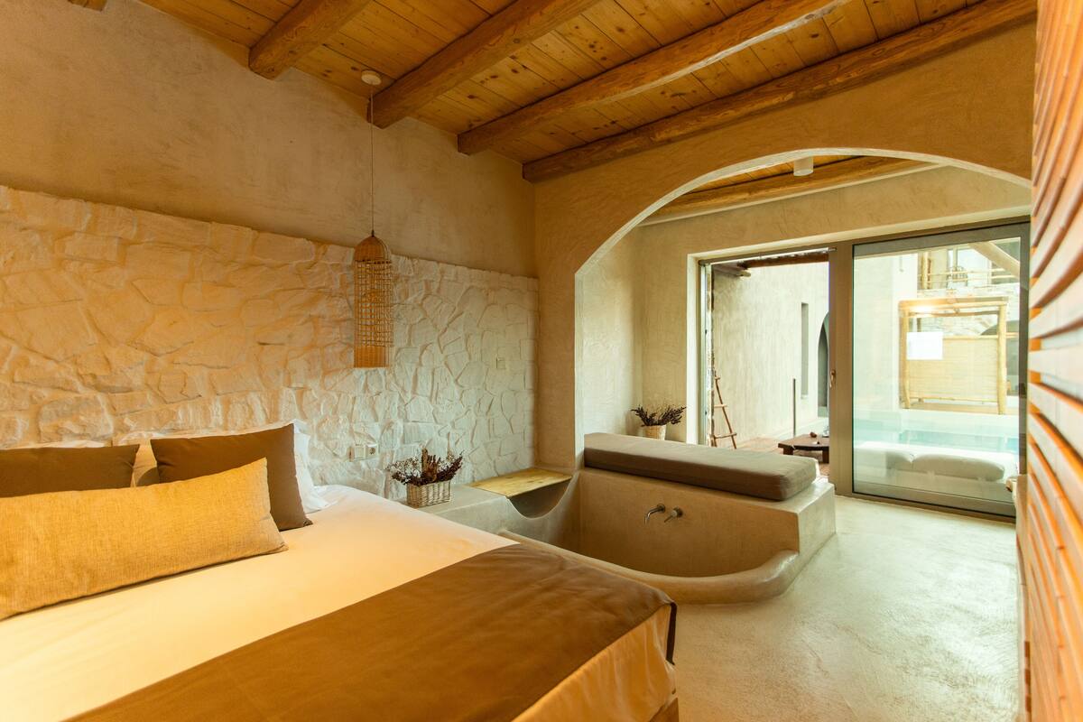 A bedroom with a stone wall, a wooden ceiling, a bed with pillows, a nightstand, and a bathtub.