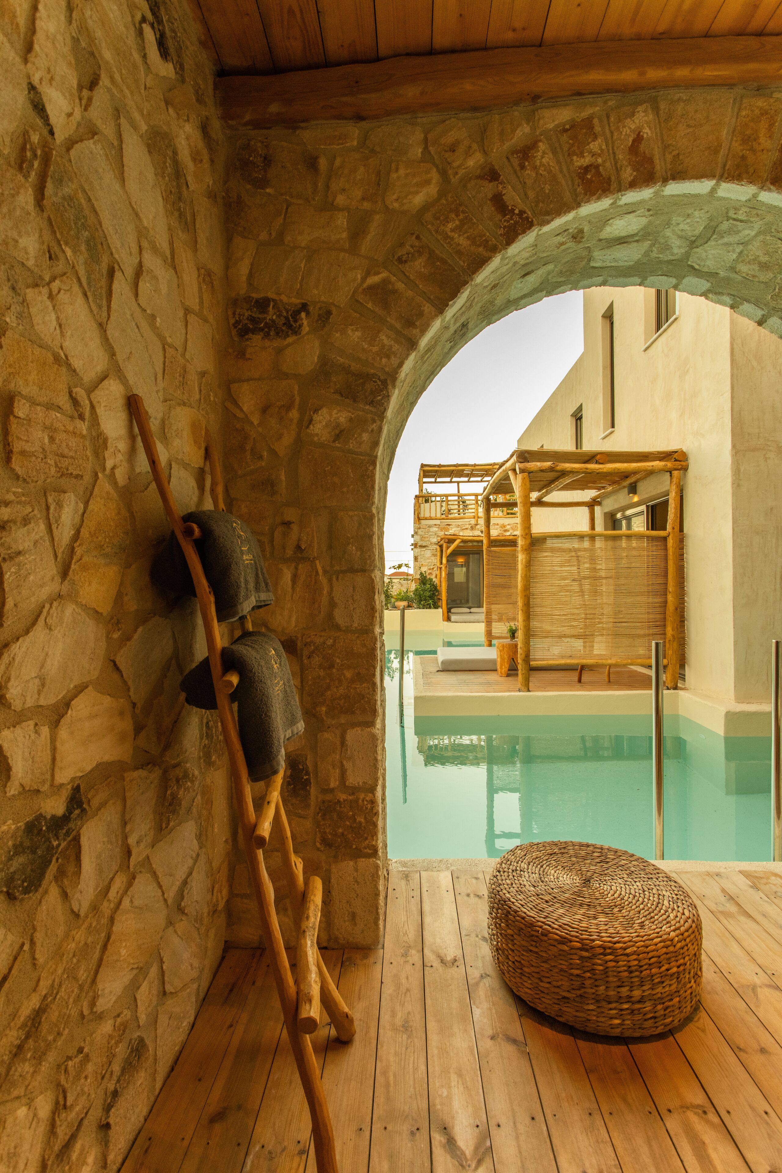 A stone-walled room with a wooden floor, a wicker ottoman, and a view of an outdoor pool area.