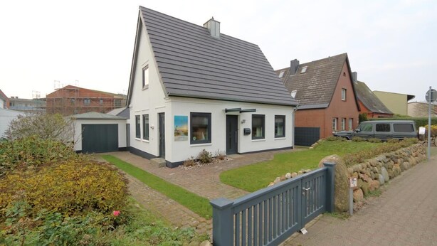 Une maison à deux étages avec un toit en pente, un garage et un jardin aménagé, agrémenté d’un mur en pierre et d’une clôture bleue.