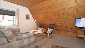 Un salon chaleureux avec un plafond en bois, un canapé, deux fauteuils, un téléviseur et une fenêtre avec des rideaux.