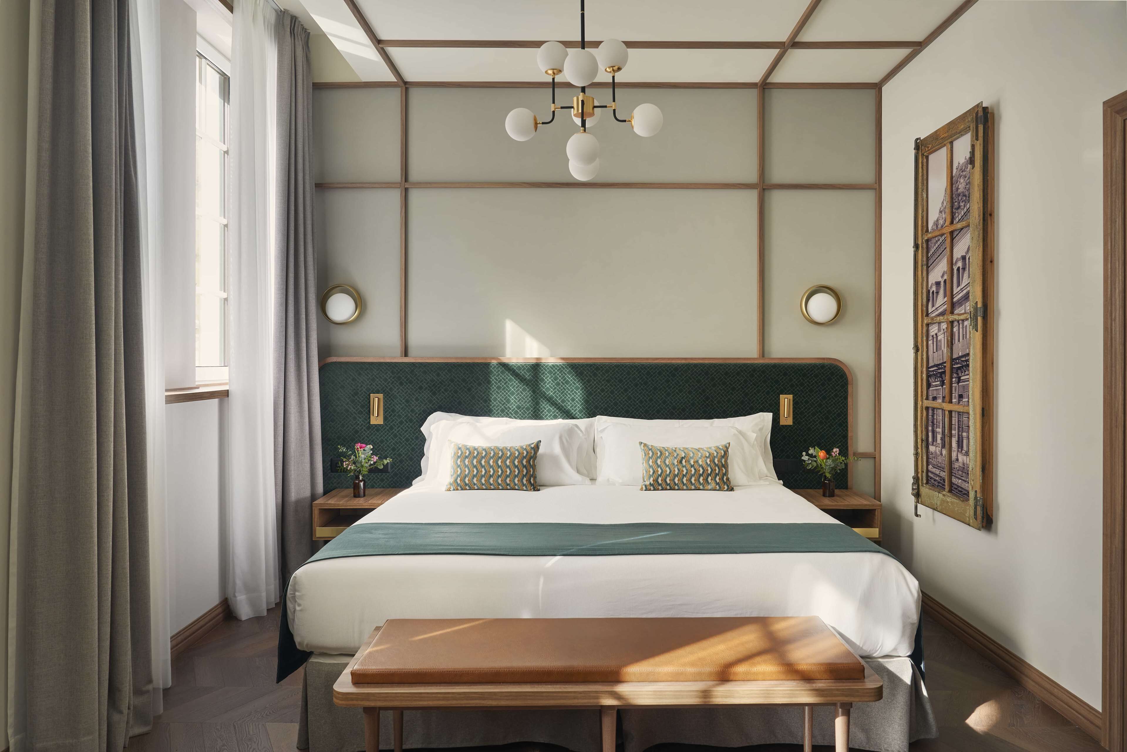 A hotel room with a large bed, bedside tables, a bench, a chandelier, and a window with curtains.