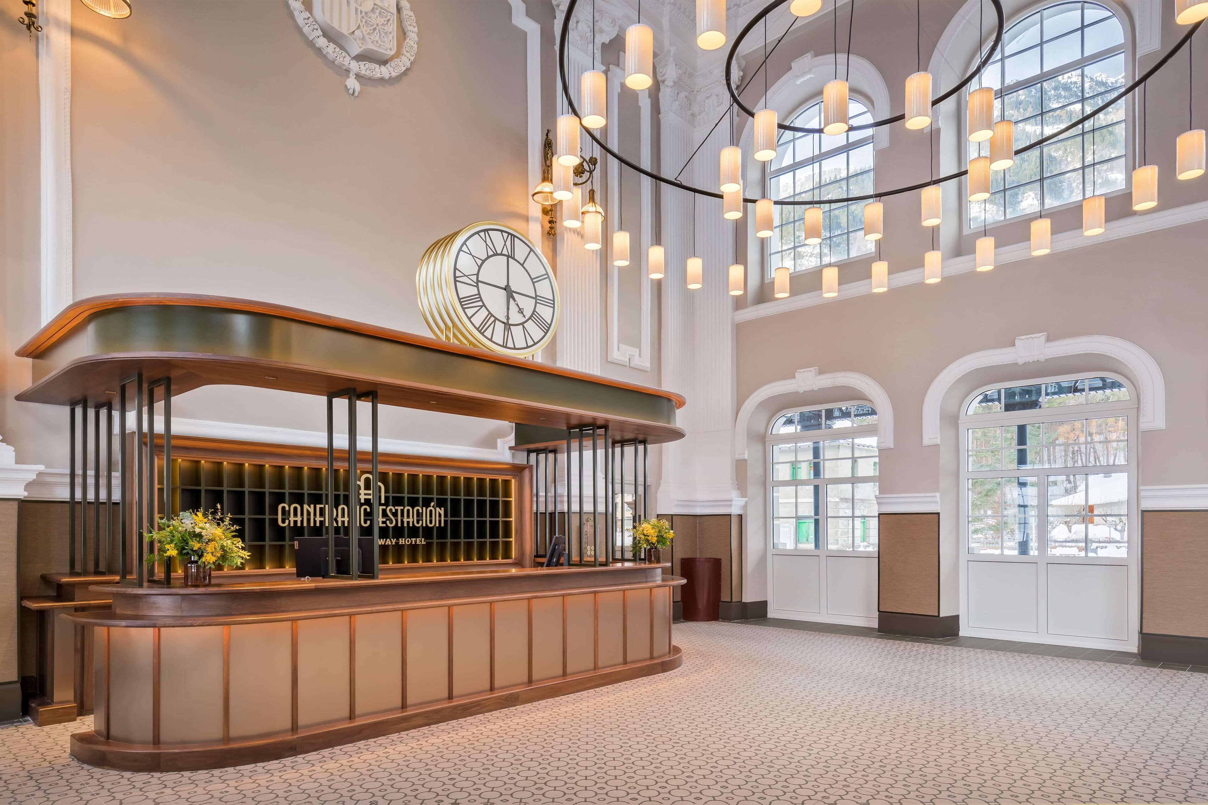 A reception desk with a clock, a chandelier, and a view of the outside through large windows.