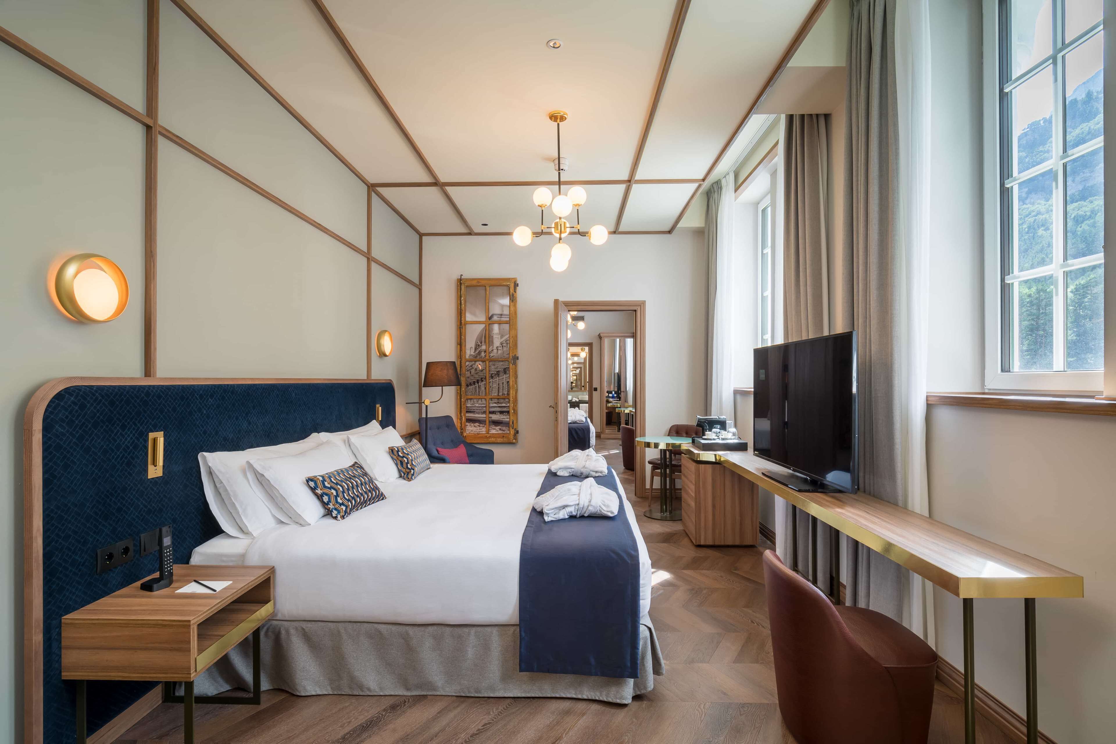 A hotel room with a large bed, a desk with a TV, and a view of the outdoors through the window.