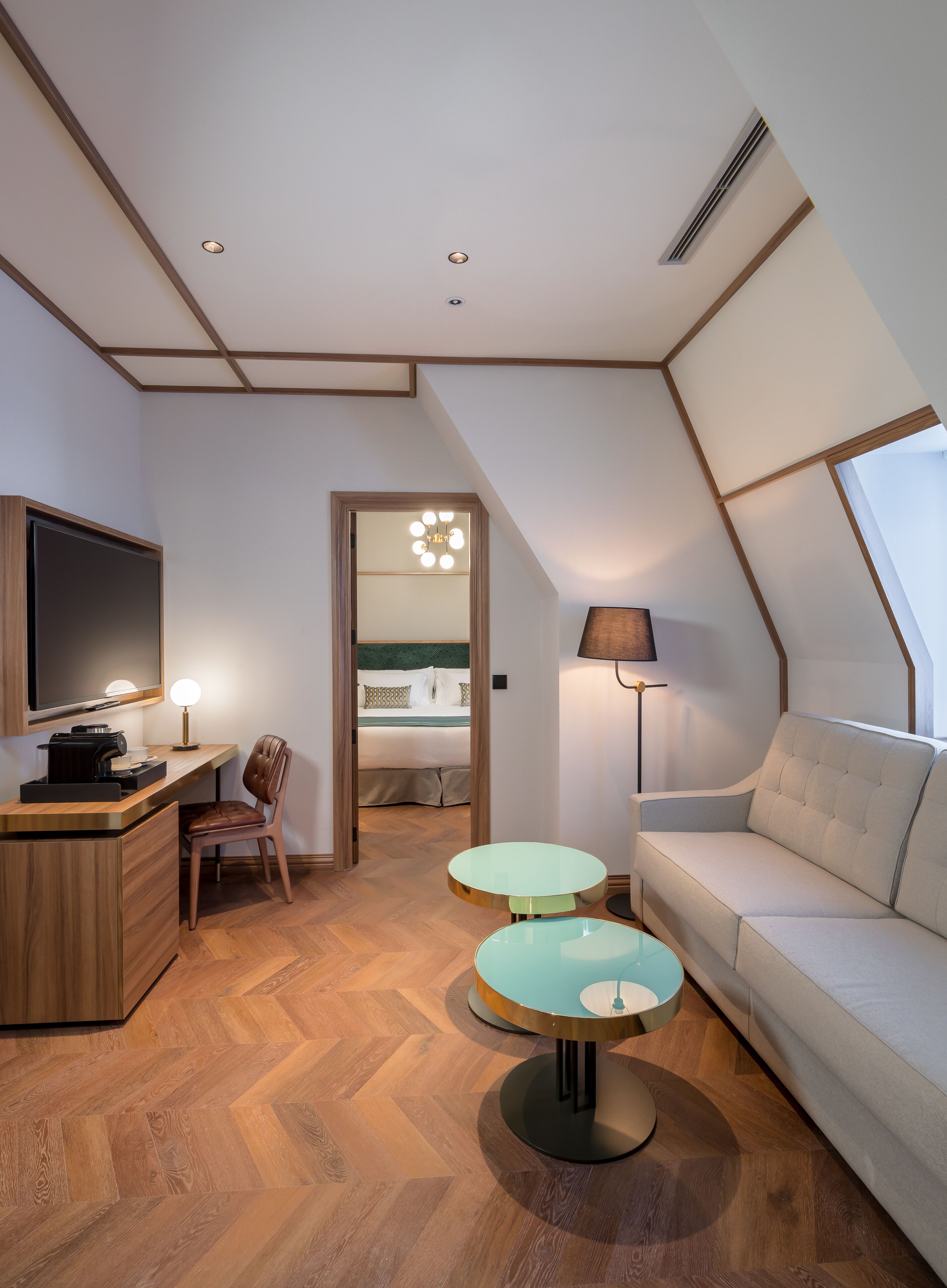 Quarto de hotel moderno com teto em estilo loft, televisão de tela plana, escrivaninha com cafeteira, sofá, duas mesinhas e cama com criado-mudo.