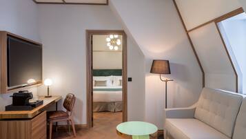 Ein modernes Hotelzimmer mit schrägedachähnlicher Decke, einem Flachbildfernseher, einem Schreibtisch mit Kaffeemaschine, einer Couch, zwei kleinen Tischen und einem Bett mit Nachttisch.