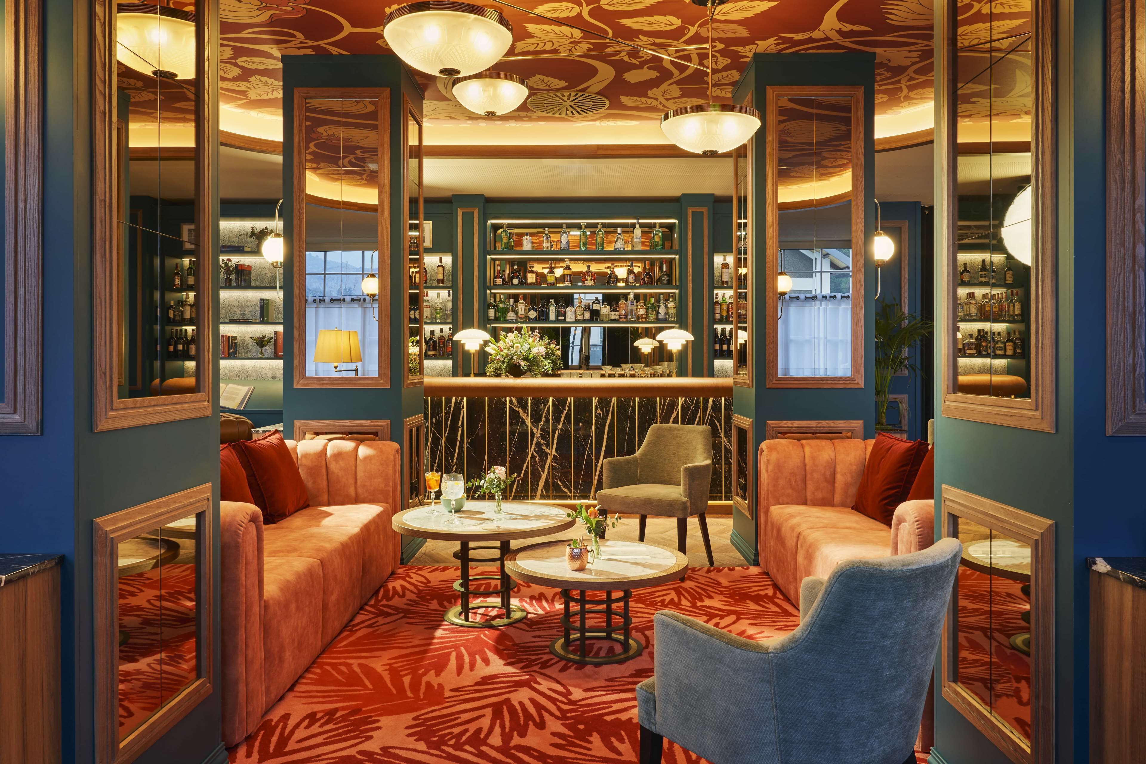 Un coin bar avec une variété de bouteilles, un canapé et un fauteuil, dans une pièce dotée d’un tapis à motifs et de quelques éléments décoratifs en bois.