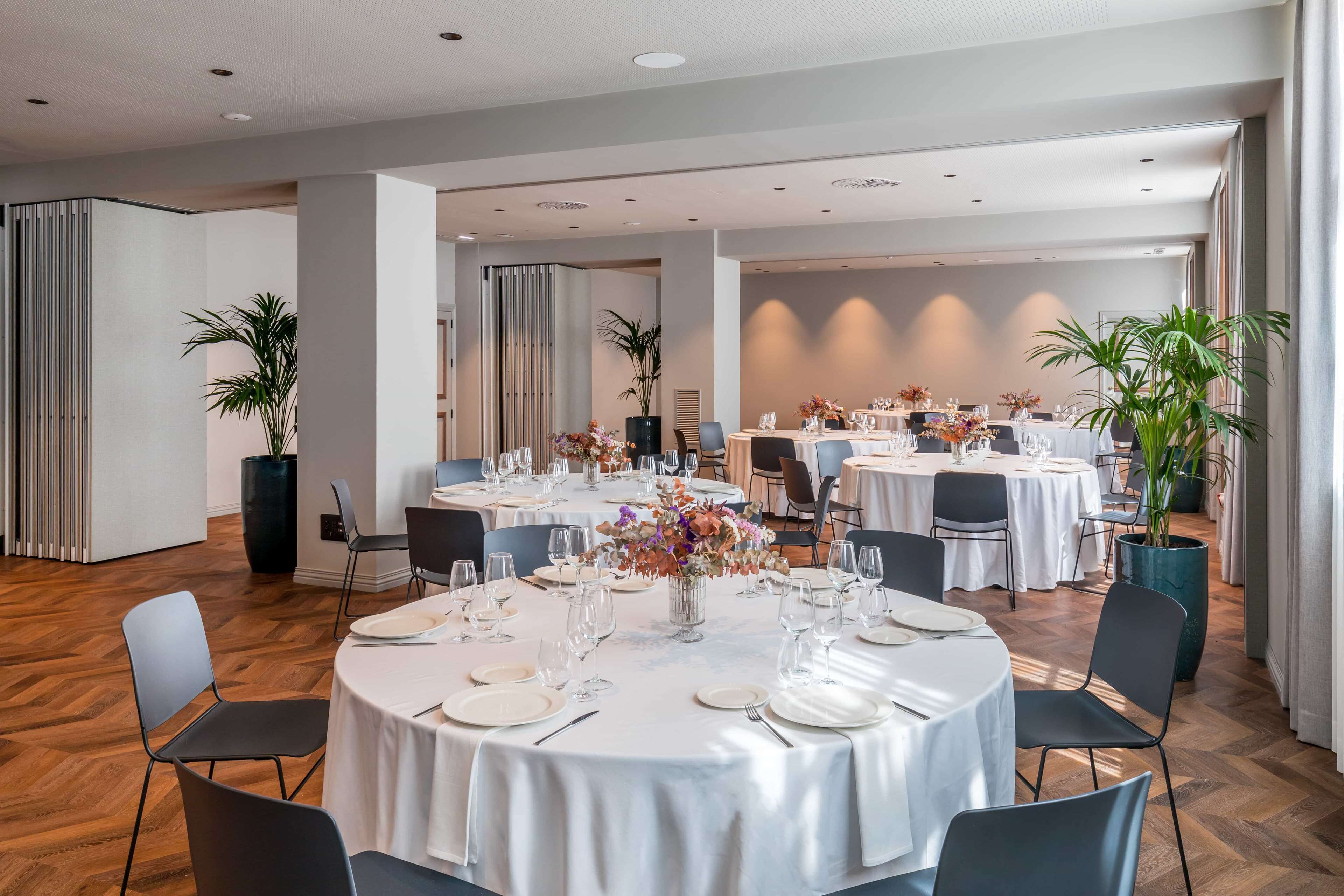 A spacious dining area with round tables set for a formal event, featuring white tablecloths and floral centerpieces.