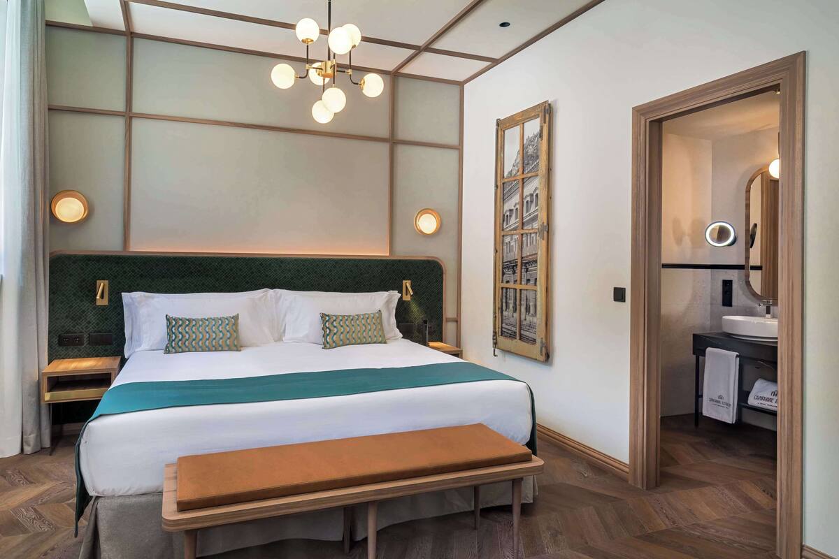 A neatly arranged hotel room with a large bed, bedside tables, a bench, a hanging light fixture, a framed picture, and a bathroom visible through an open door.