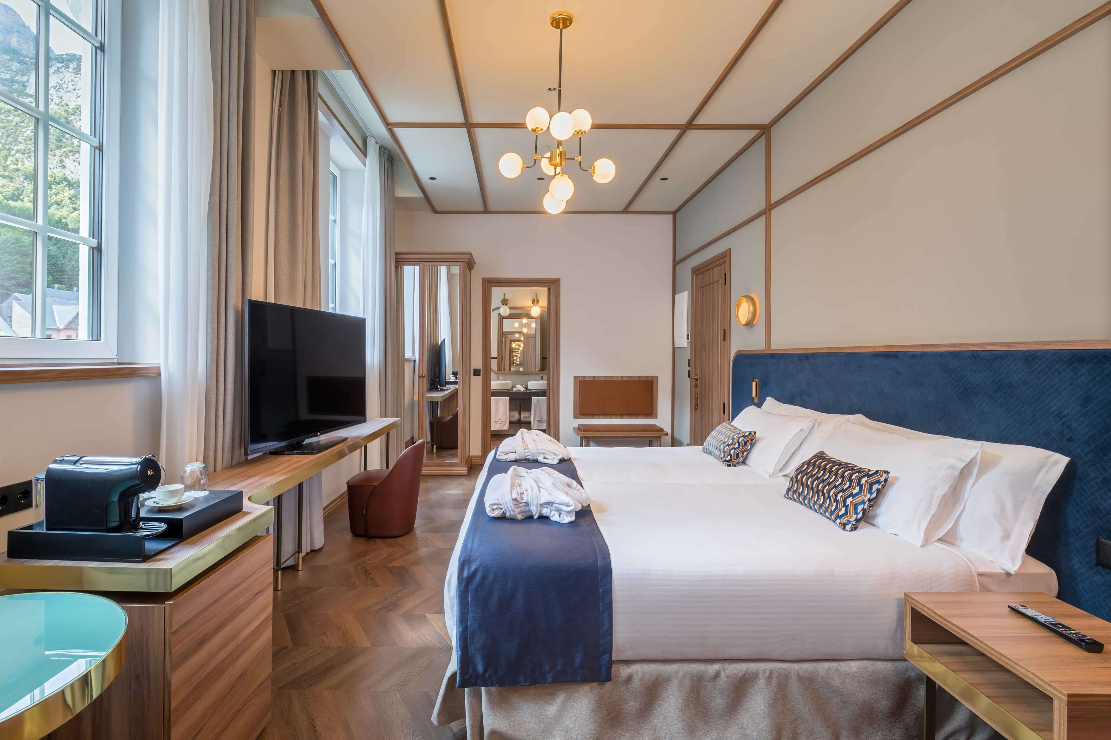 Habitación de hotel con una cama grande, televisión, escritorio con cafetera y vistas al exterior a través de amplias ventanas.