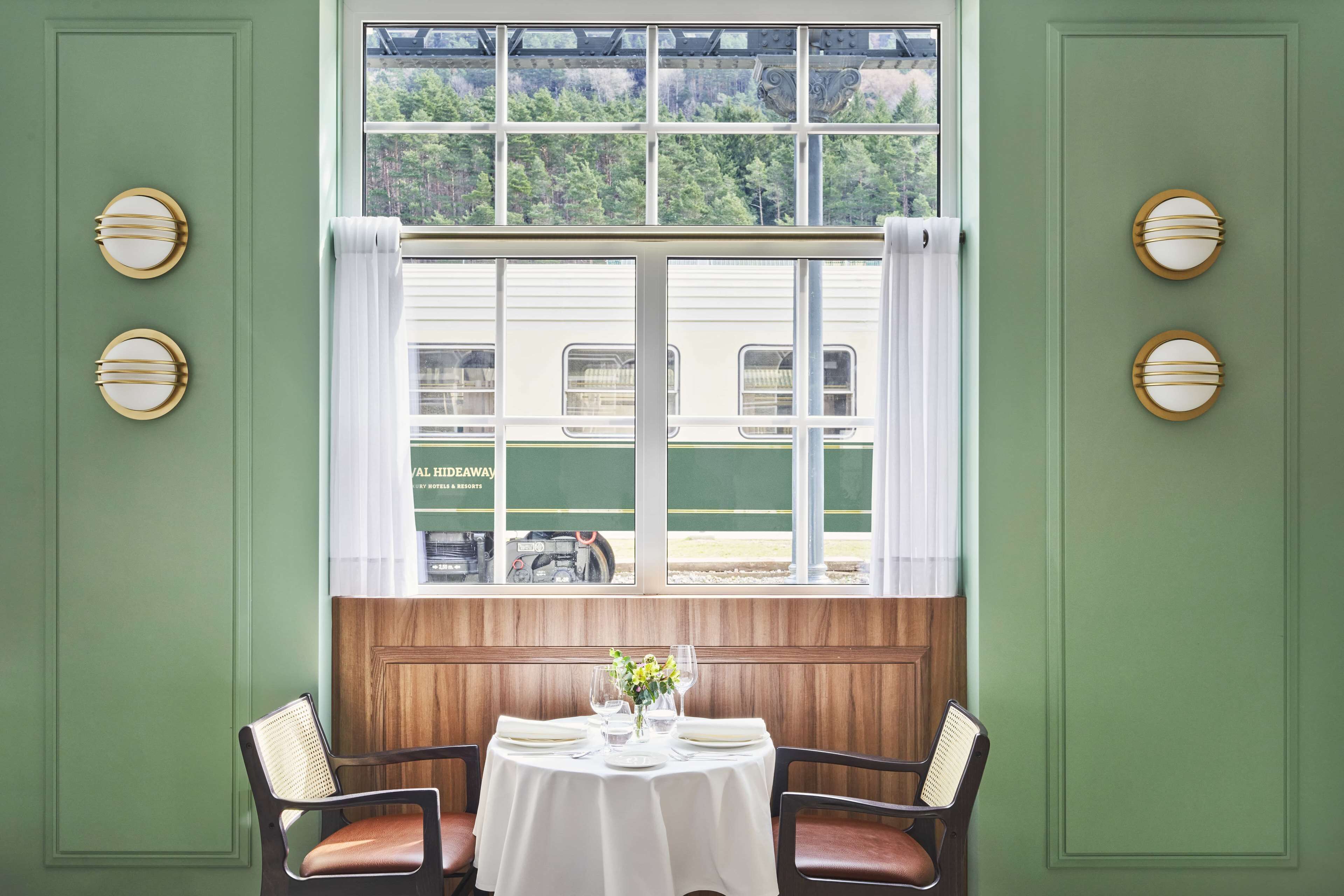 Un comedor con una mesa dispuesta para dos, una ventana con vista a un tren y una pared verde con detalles circulares decorativos.