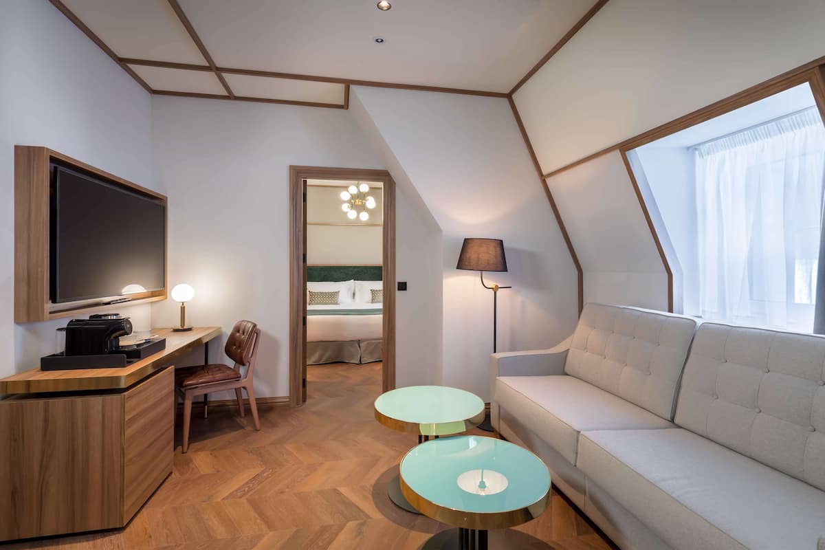 A modern hotel room with a flat-screen TV, a desk with a coffee machine, two round tables, a sofa, and a large window with curtains.