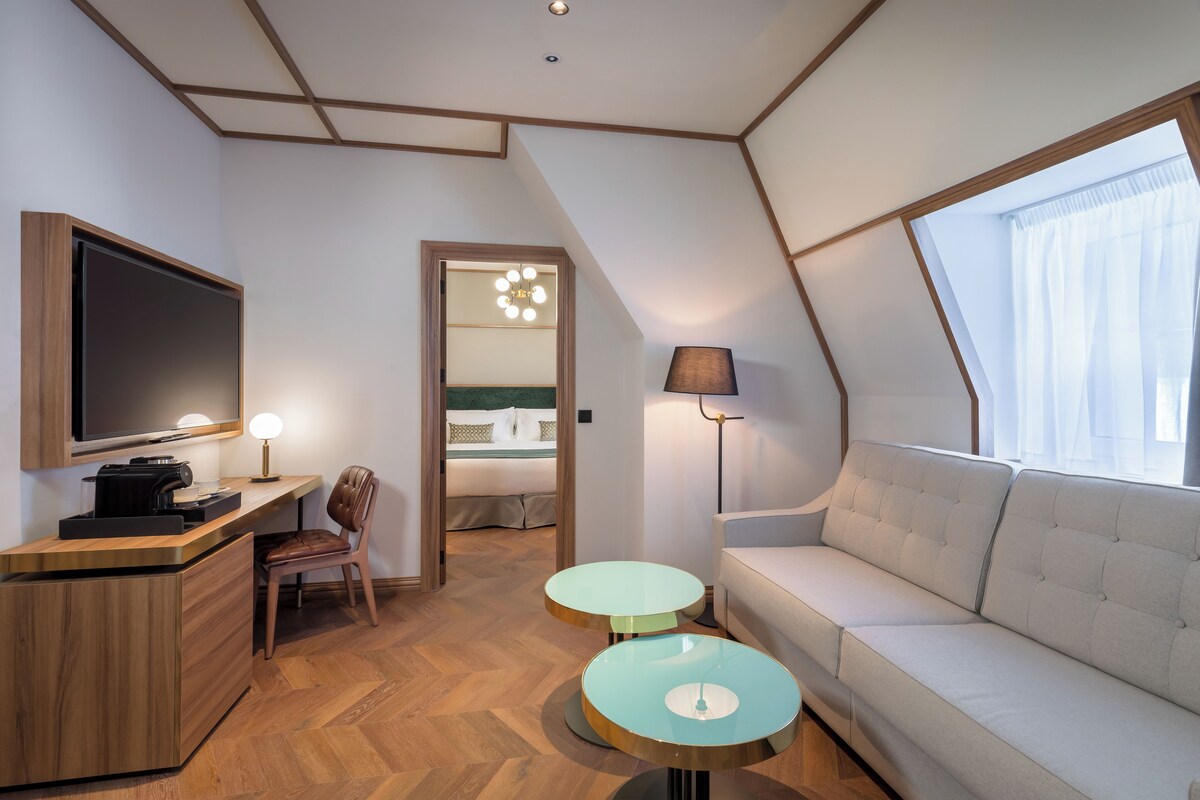 Una habitación de hotel moderna con una televisión de pantalla plana, un sofá, dos mesitas, una silla y un amplio ventanal con cortinas.