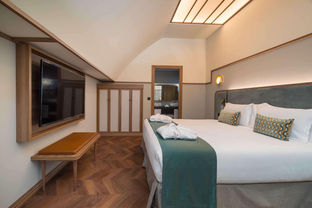 Una habitación de hotel con cama, televisor montado en la pared, una banca y un espejo.