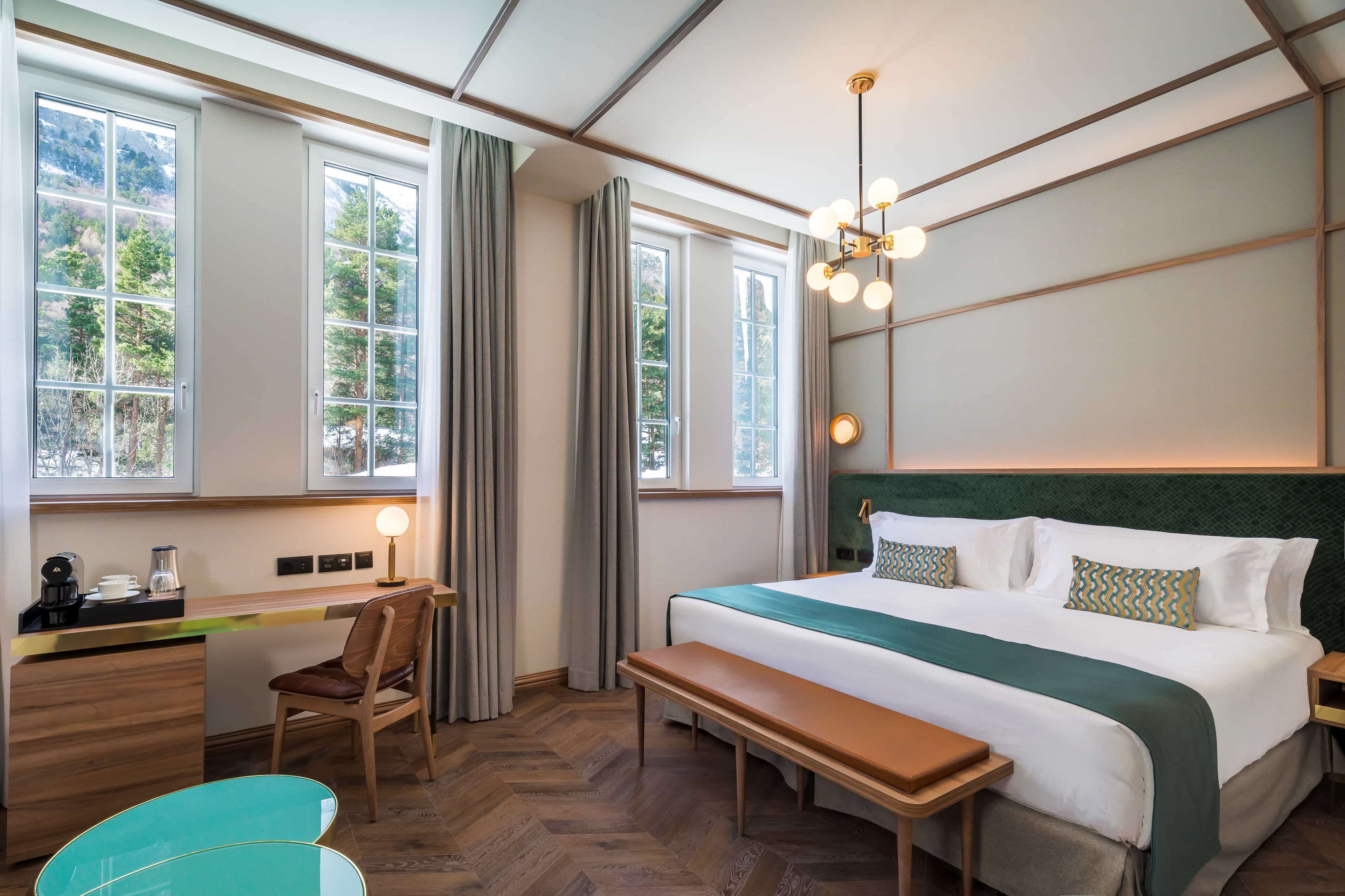 A hotel room with a large bed, a desk with a chair, a bench, and a view of trees through the windows.