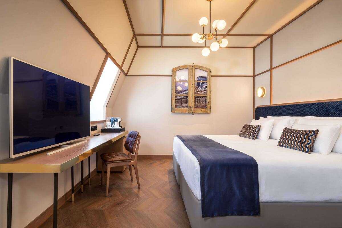 A modern hotel room with a large flat-screen TV, a desk with a chair, a bed with a dark blue blanket, and a unique wall-mounted mirror.
