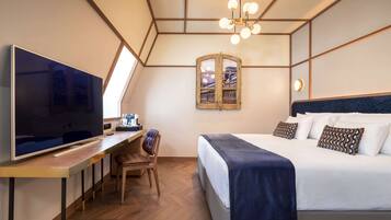 Una habitación de hotel moderna con un televisor de pantalla plana grande, un escritorio con una silla, una cama con una manta azul oscuro y un espejo original montado en la pared.