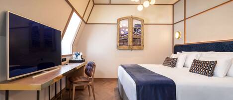 A modern hotel room with a large flat-screen TV, a desk with a chair, a bed with a dark blue blanket, and a unique wall-mounted mirror.