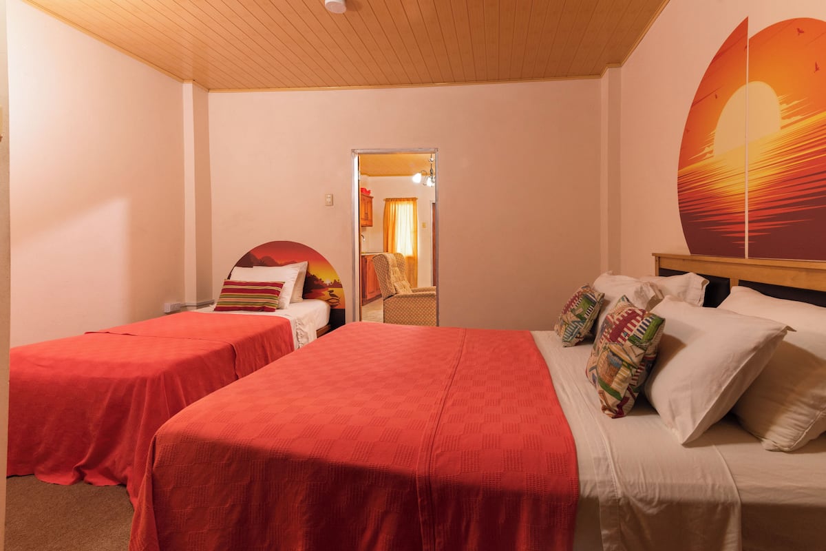 A hotel room with two beds, one with red bedding and the other with white bedding, a large window with a sunset view, and a wooden ceiling.