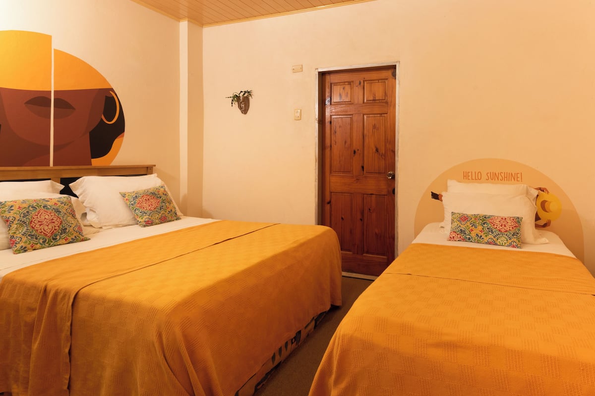 A hotel room with two beds, each with a yellow bedspread and matching pillows. There is a wooden door with a sign above it that reads "HELLO SUNSHINE!"