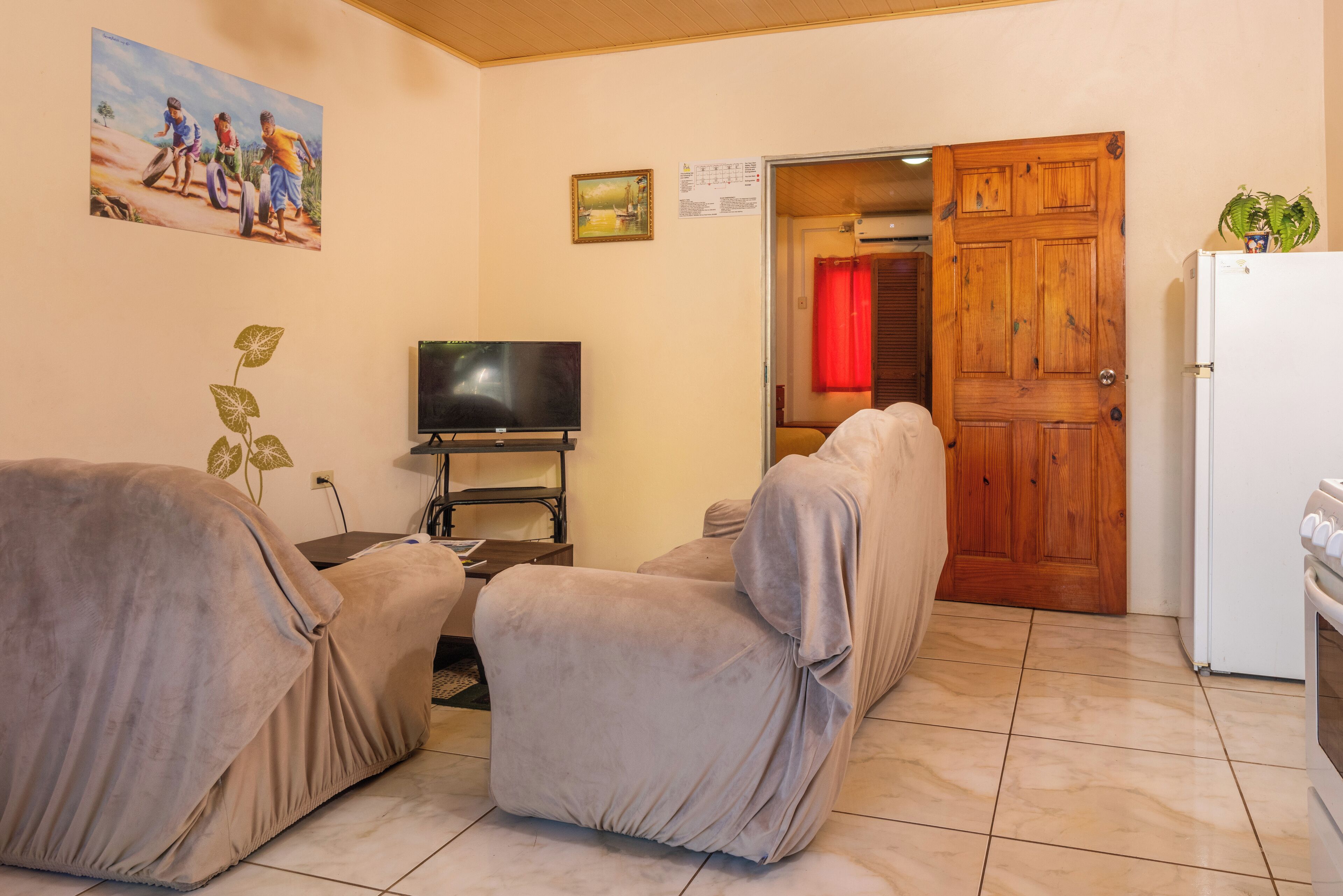A living room with two covered sofas, a television, a wooden door, and a refrigerator.