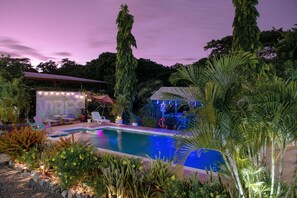 A swimming pool surrounded by tropical plants and a covered area with string lights.