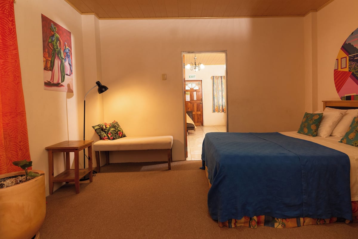 A bedroom with a bed, bench, and a small table. There is a door leading to another room.