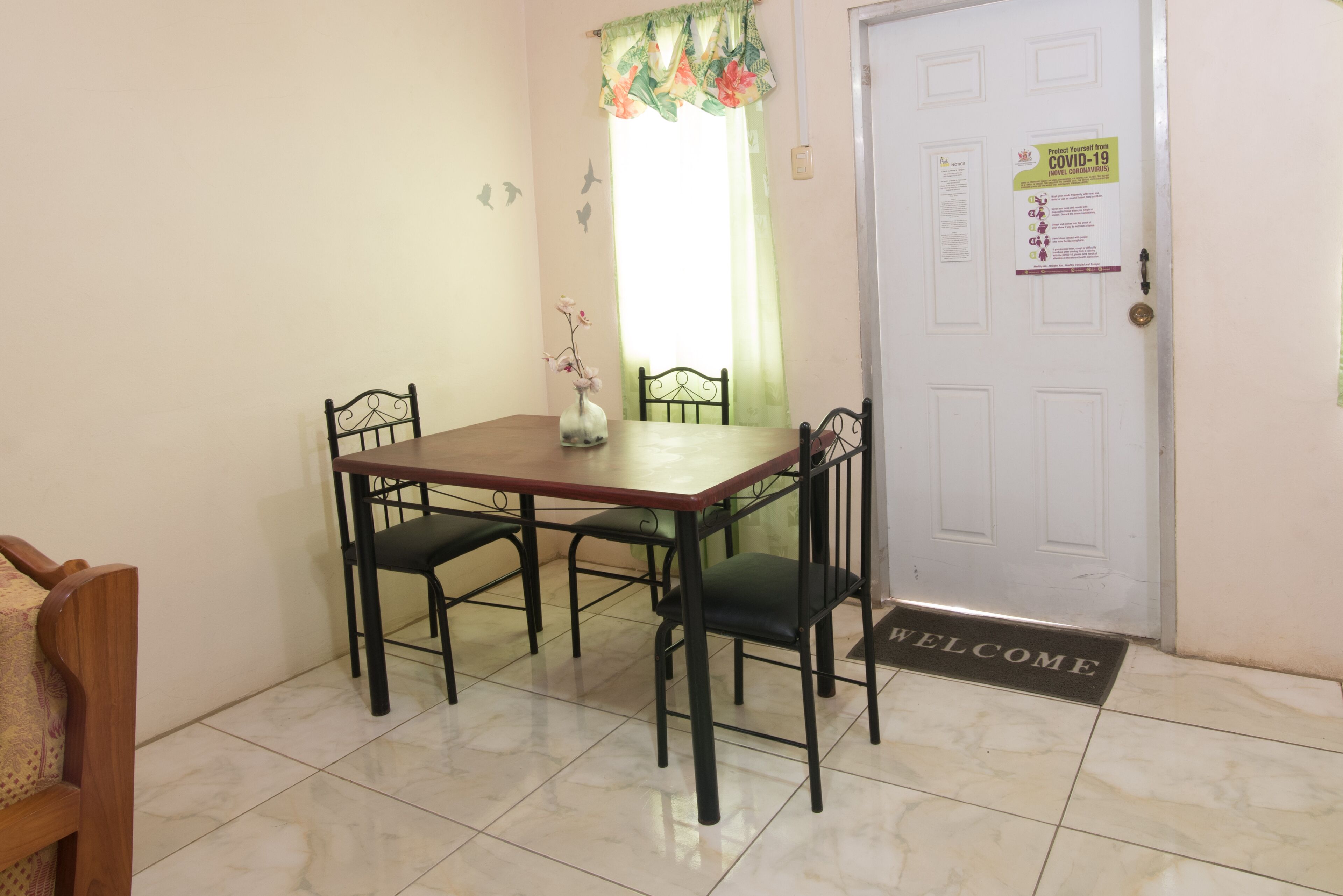 A dining area with a wooden table and four chairs, a vase with flowers, a door with a 'WELCOME' mat, and a COVID-19 informational sign.