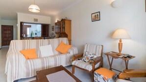 Una sala de estar con un sofá a rayas, sillones, una mesita de madera con una lámpara y una cocina al fondo.