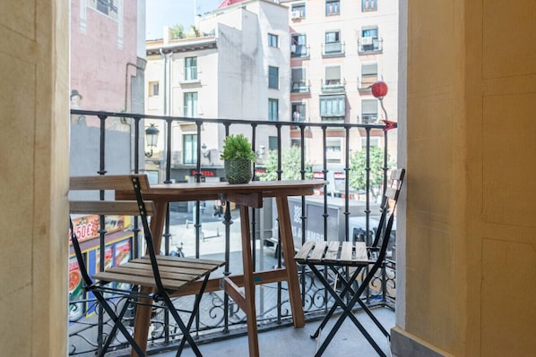 A balcony with two wooden chairs and a small table, a potted plant, and a view of a city street with buildings and a red spherical object.