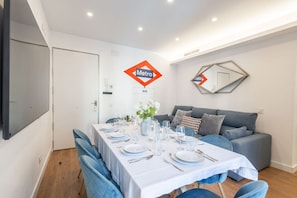 A modern dining area with a white table set for four, blue chairs, and a grey sofa in the background.