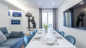 A dining area with a long table set for six, blue chairs, a white tablecloth, and a cityscape view through large windows. - 1)The Heart Of The City, Sol, By MoNARO (Madrid)