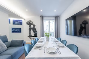 Un comedor con una larga mesa dispuesta para seis comensales, sillas azules, un mantel blanco y vistas a la ciudad a través de amplios ventanales.