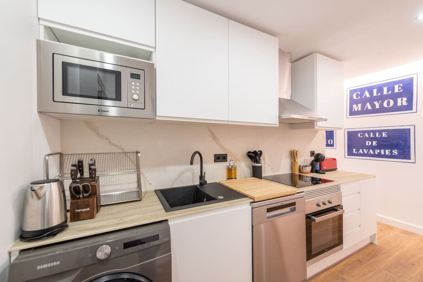 Een moderne keuken met een magnetron, spoelbak en vaatwasser. Er zijn kastjes, een snijplank en diverse keukengerei.