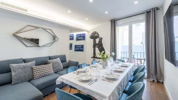 A modern dining area with a long table set for six, a grey sofa, and a decorative wall piece. - 1)The Heart Of The City, Sol, By MoNARO (Madrid)