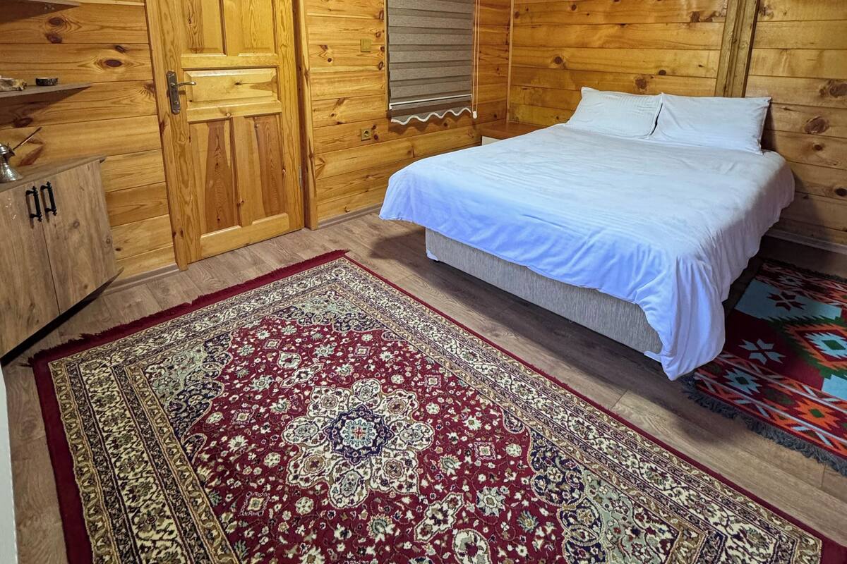 A wooden cabin bedroom with a bed, a red patterned rug, a wooden dresser, and a door.