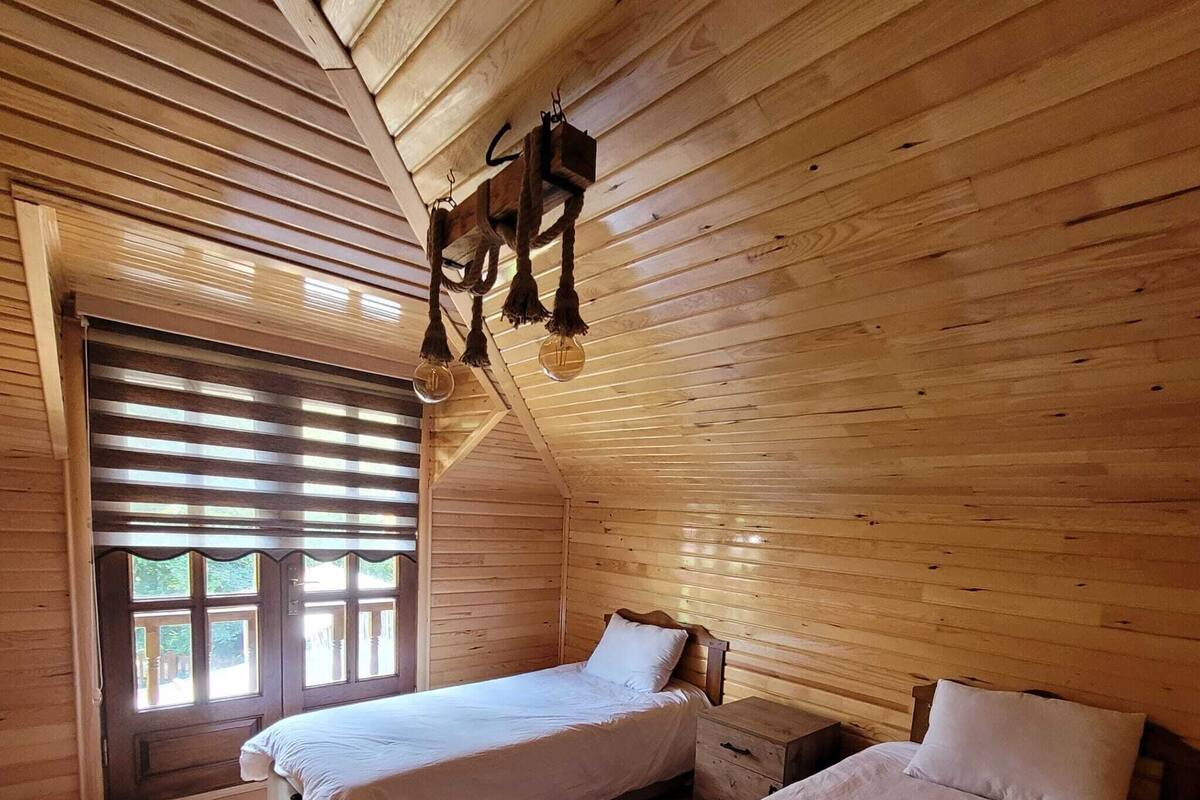 A cabin-style bedroom with two beds, a window with blinds, and a hanging light fixture.