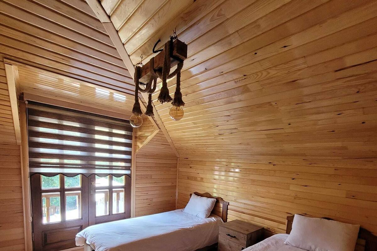 A cozy cabin-style bedroom with two single beds, a small wooden nightstand, and a large window with blinds.