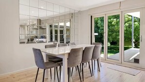 A modern dining area with a white table and grey chairs, large windows, and a view of greenery outside.