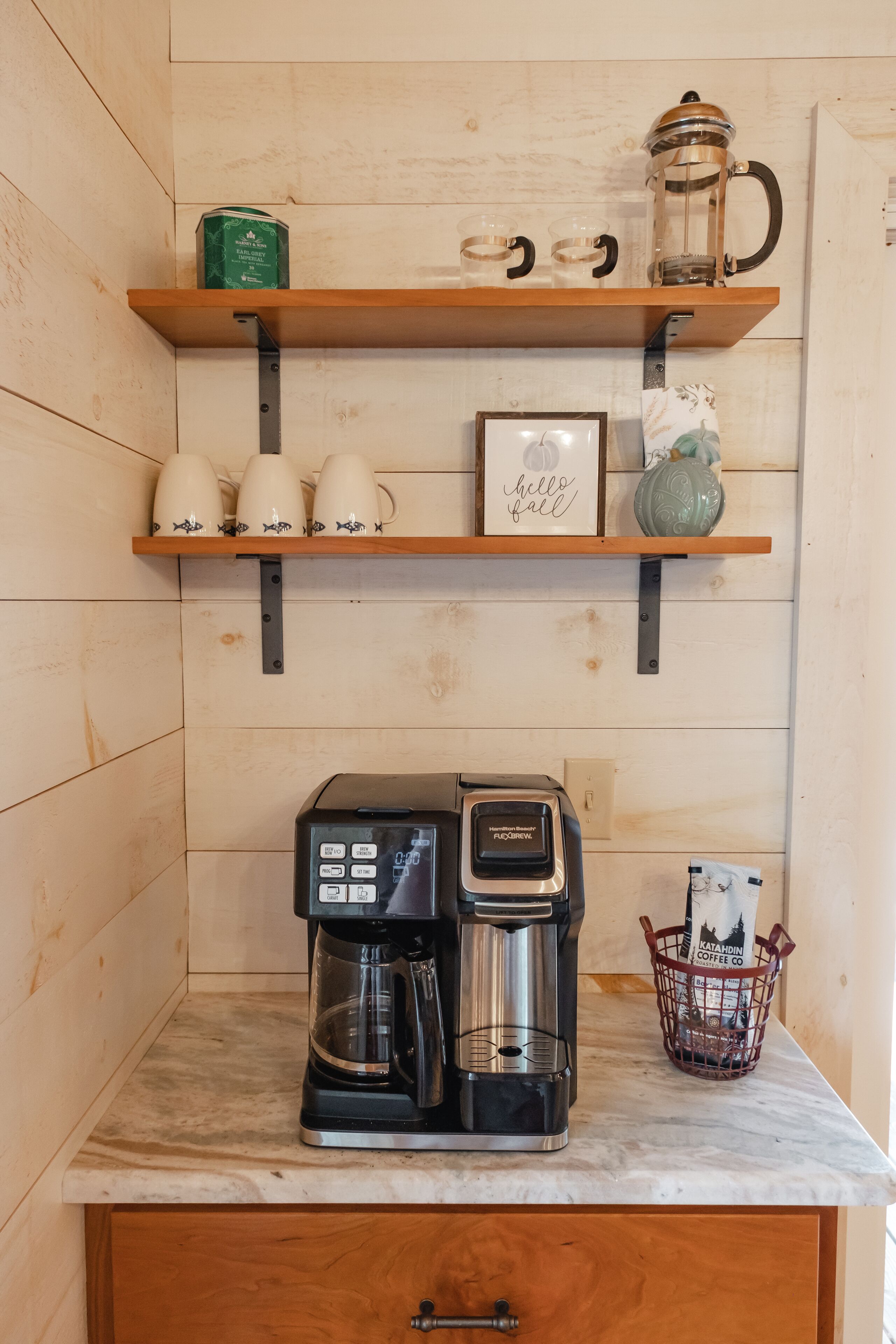 A coffee station with a coffee maker, cups, and a basket of coffee pods.