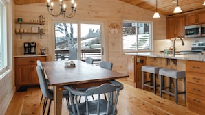 L’intérieur d’un chalet en bois comprend une table à manger, des chaises, des tabourets de bar, un coin cuisine et une grande fenêtre donnant sur un paysage enneigé.