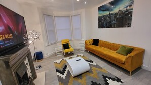 A modern living room with a large flat-screen TV, a yellow sofa, a patterned rug, and a cityscape painting on the wall.
