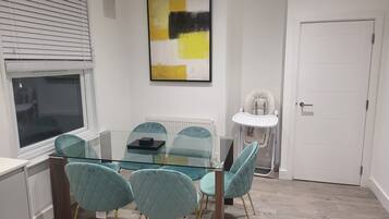 A dining area with a glass table and teal chairs, a white radiator, a baby high chair, and a door.