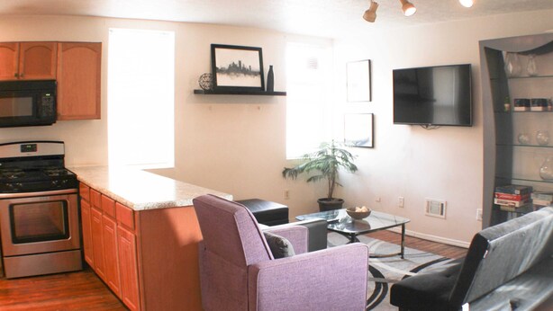 A modern kitchen with wooden cabinets, a microwave, and a stove. There is a dining area with a table and chairs, a TV mounted on the wall, and a potted plant. - Carey Way Quarters-Stylish 3BR Loft-Prime Location (Pittsburgh)